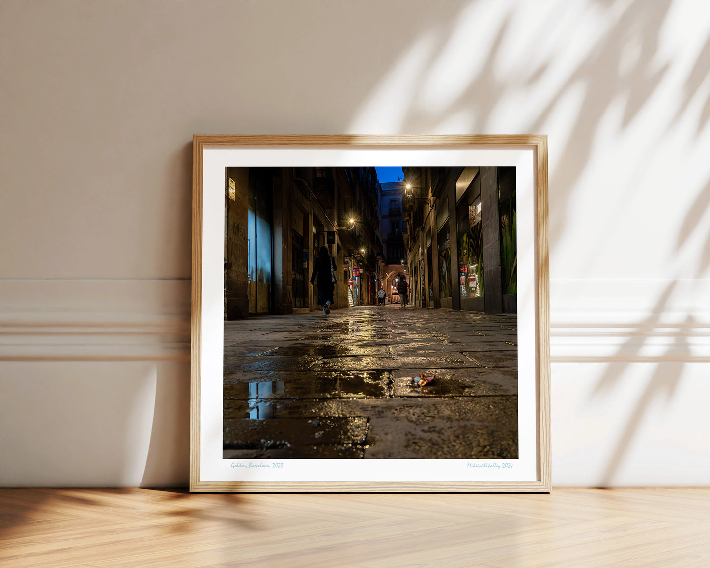 Gasse in Barcelona. Blaue Stunde. Wasserpfütze im Vordergrund. Mehrere Laternen mit Lichterkranz. Vereinzelte Passanten.