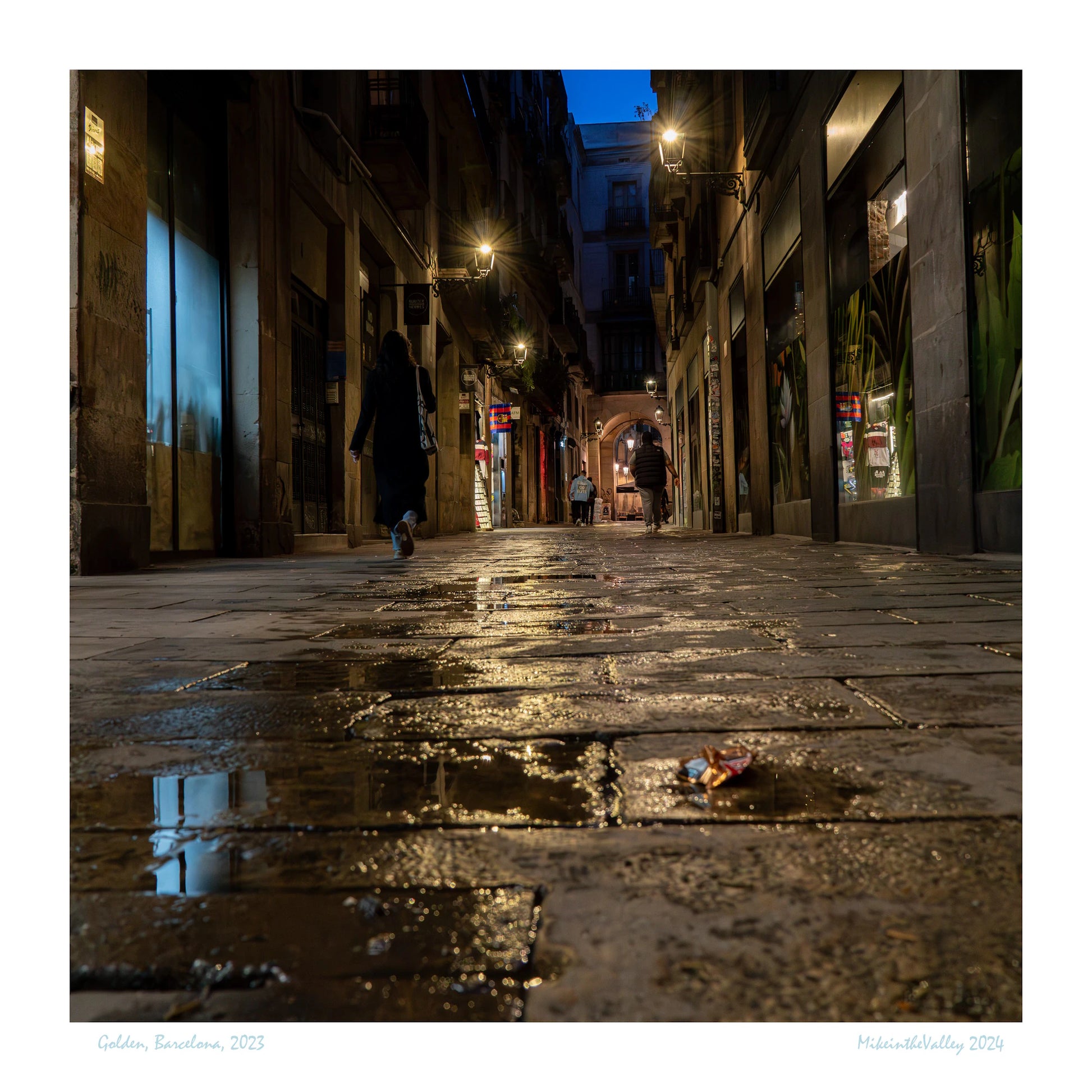 Gasse in Barcelona. Blaue Stunde. Wasserpfütze im Vordergrund. Mehrere Laternen mit Lichterkranz. Vereinzelte Passanten.