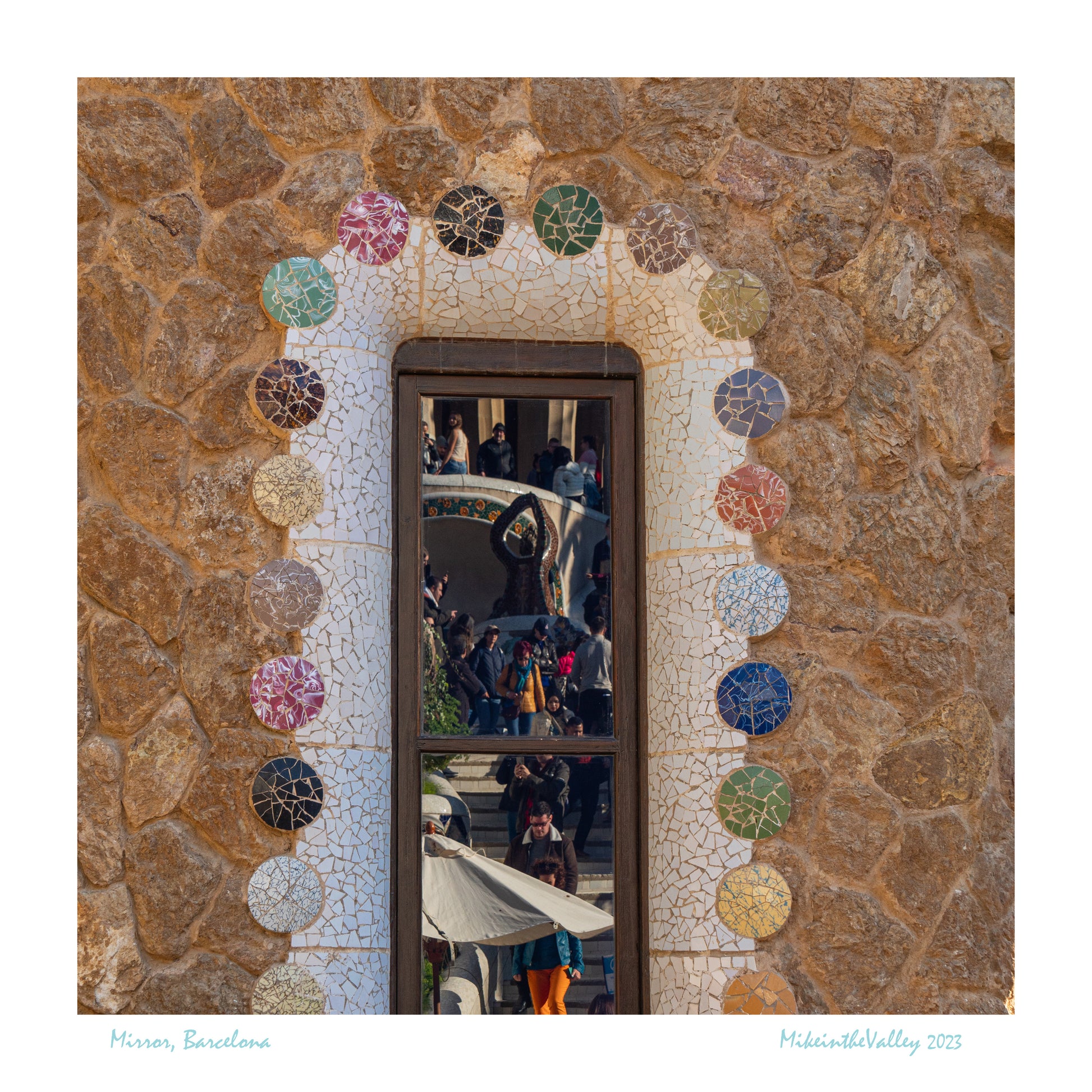 Hausfassade mit Fenster in Barcelona. Fenster ist mit bunten Keramik-Teilen in Trencadis-Technik eingefasst. Im Fenster spiegeln sich Menschen, die eine Treppe hinuntergehen.