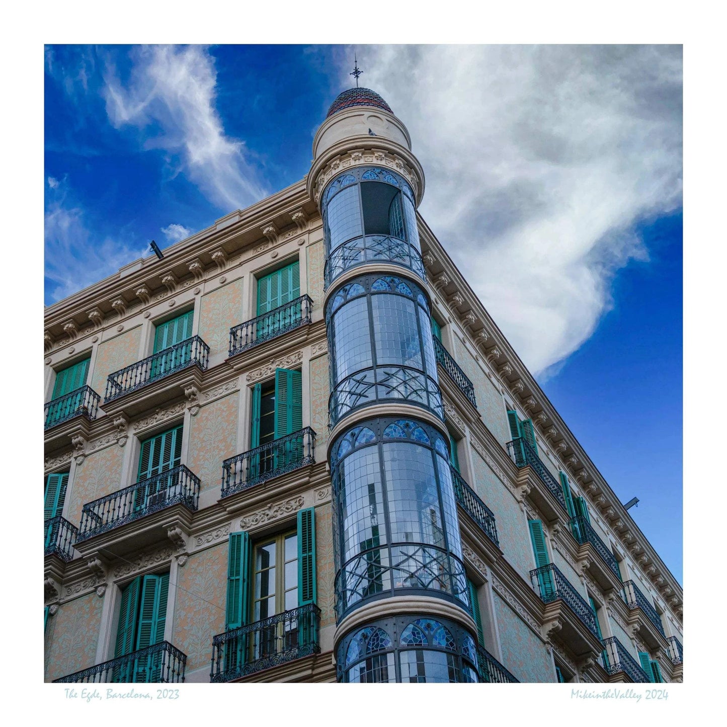 Ein Eckhaus in Barcelona, das Casas Torre Andreu. Grüne Fensterläden und ein runder, blau verglaster Eckturm vor blauem Himmel mit Wolken.