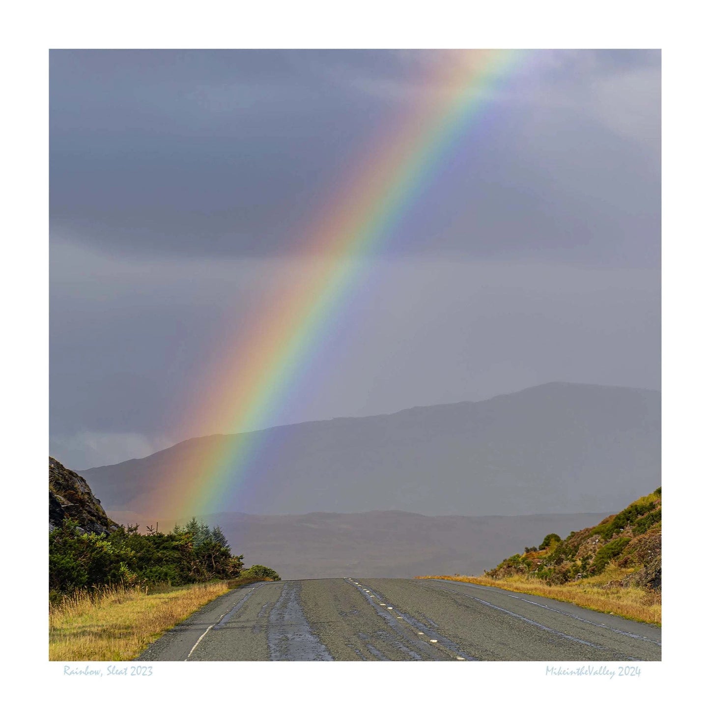 Ein kräftiger Regenbogen erhebt sich über eine Straße im Süden der Insel Skye in Schottland. Im Hintergrund eine Bergkette.