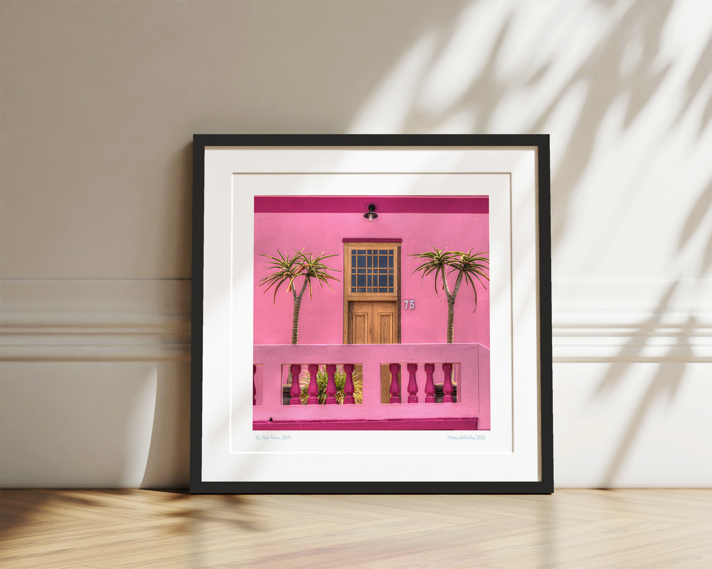 schöne Haustür aus Holz in einer pinkfarbenen Hauswand. Rechts und links davon jeweils eine Palme. Schwarzer Rahmen mit Passepartout.